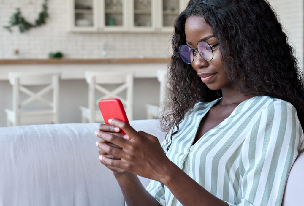 Young happy smart African black student girl businesswoman in glasses sitting on sofa holding using cell mobile phone apps reading e book, surfing social media, online chatting in apartment.